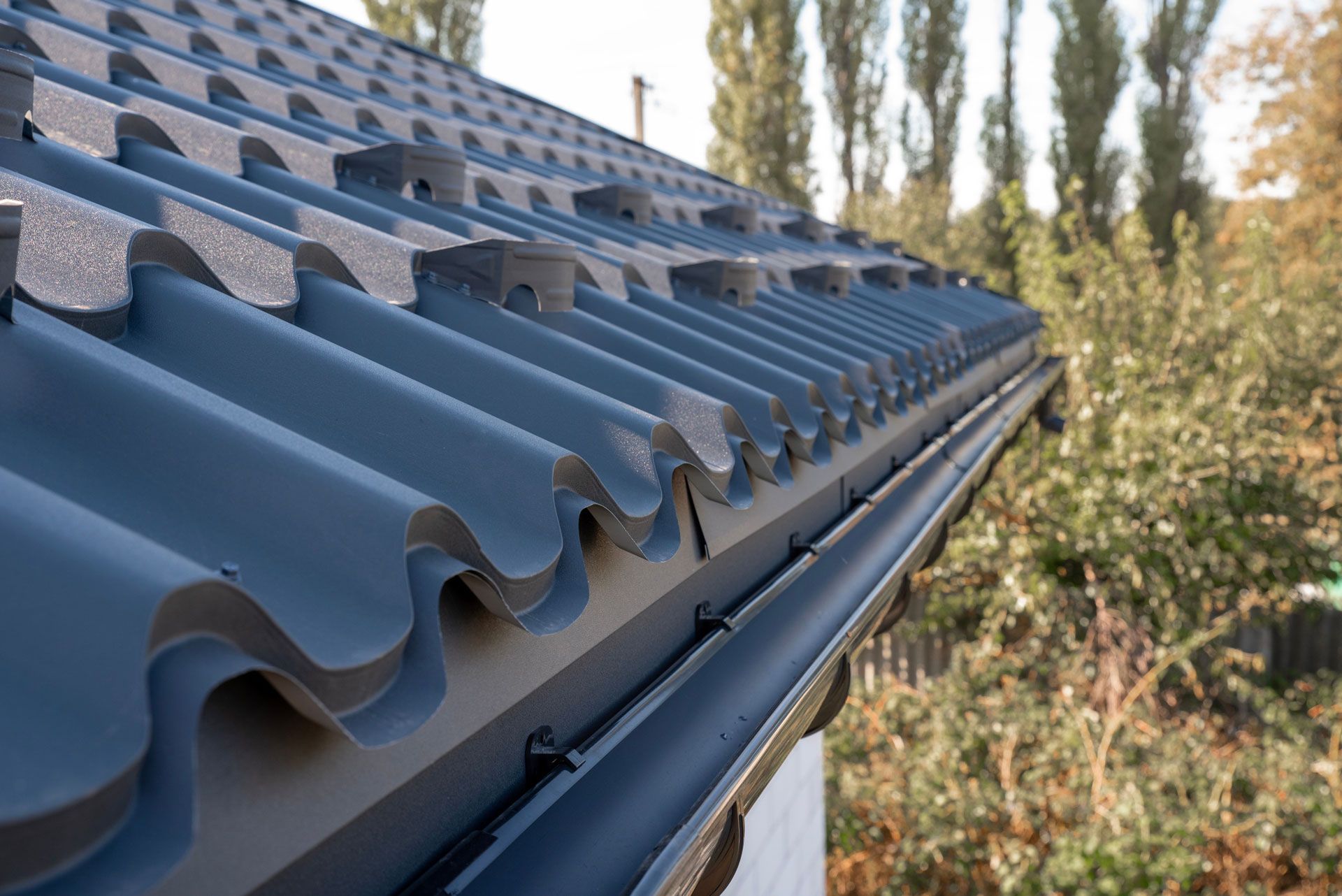 A close-up of dark grey, corrugated metal roof panels installed over a matching gutter system against a backdrop of trees.