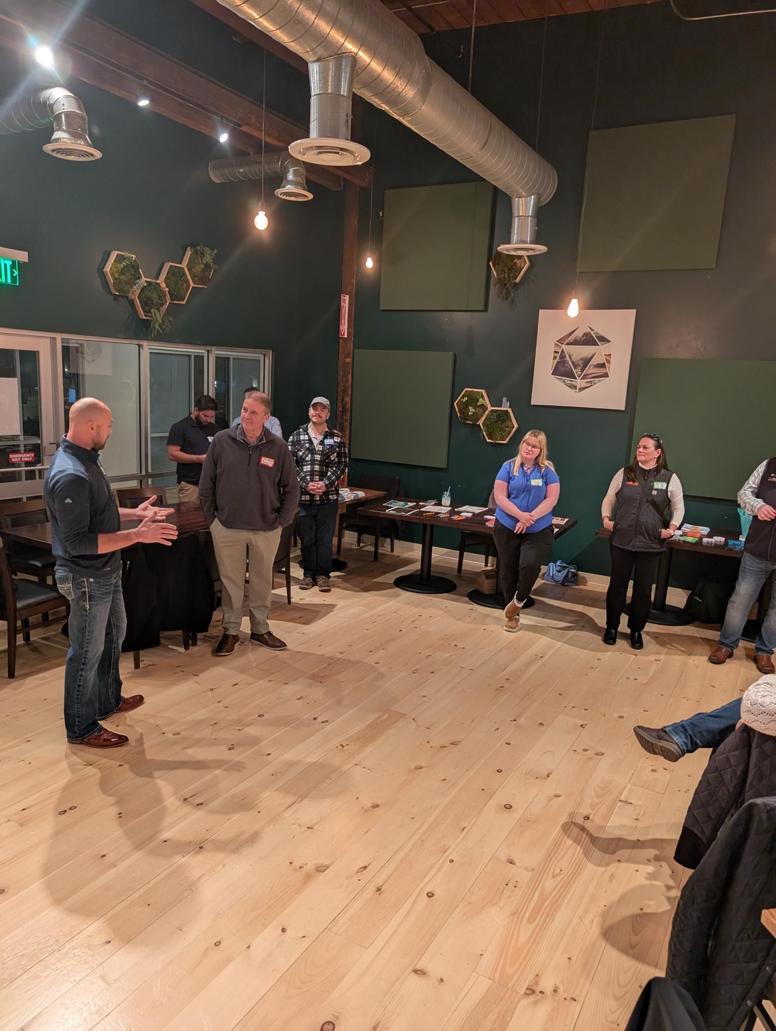 A man speaks to a group in a room. Others stand, listen. Wooden floors, dark walls, some decor.