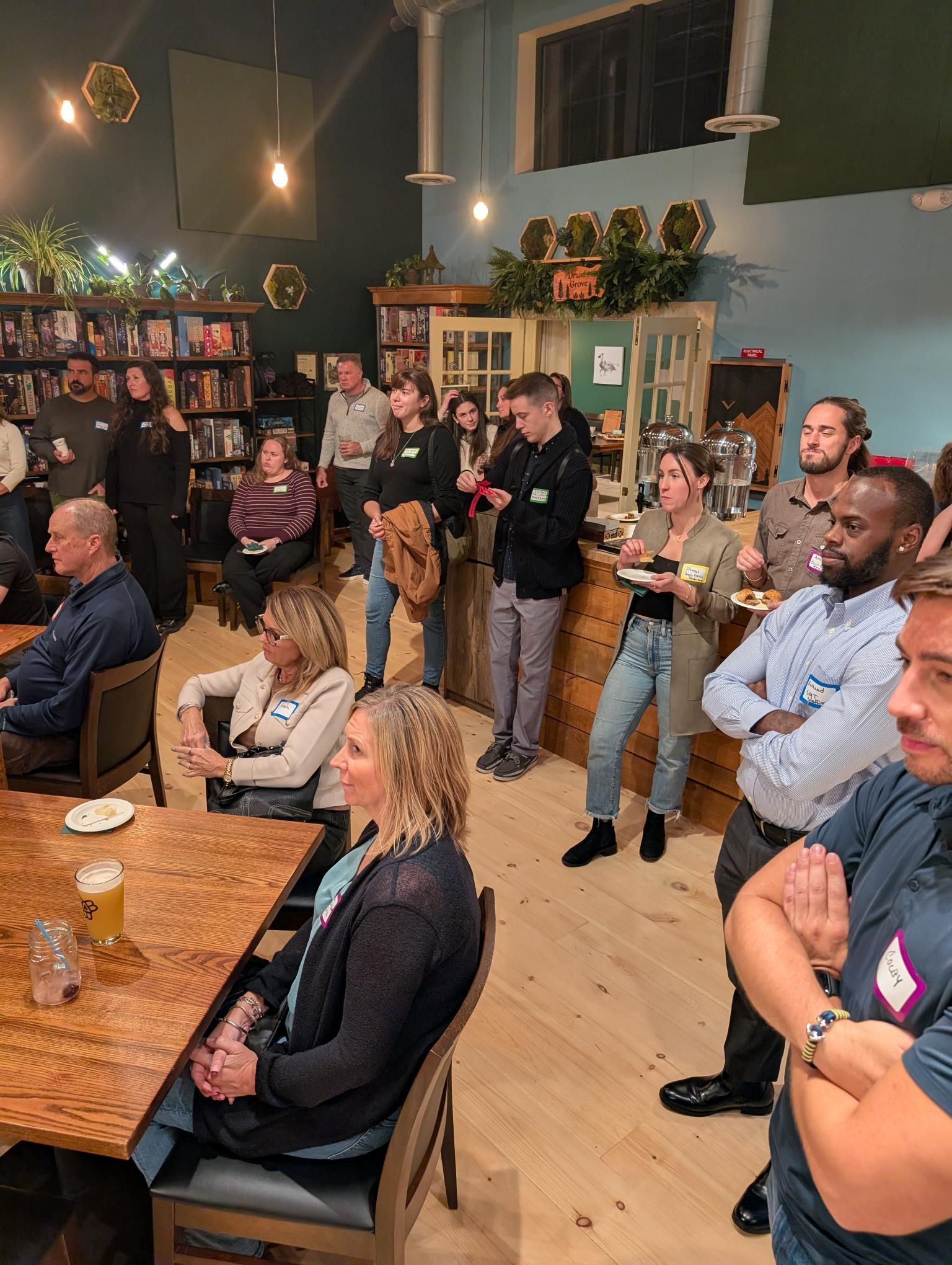 Group of people at an event in a venue with wooden tables, light wood flooring, and shelves.
