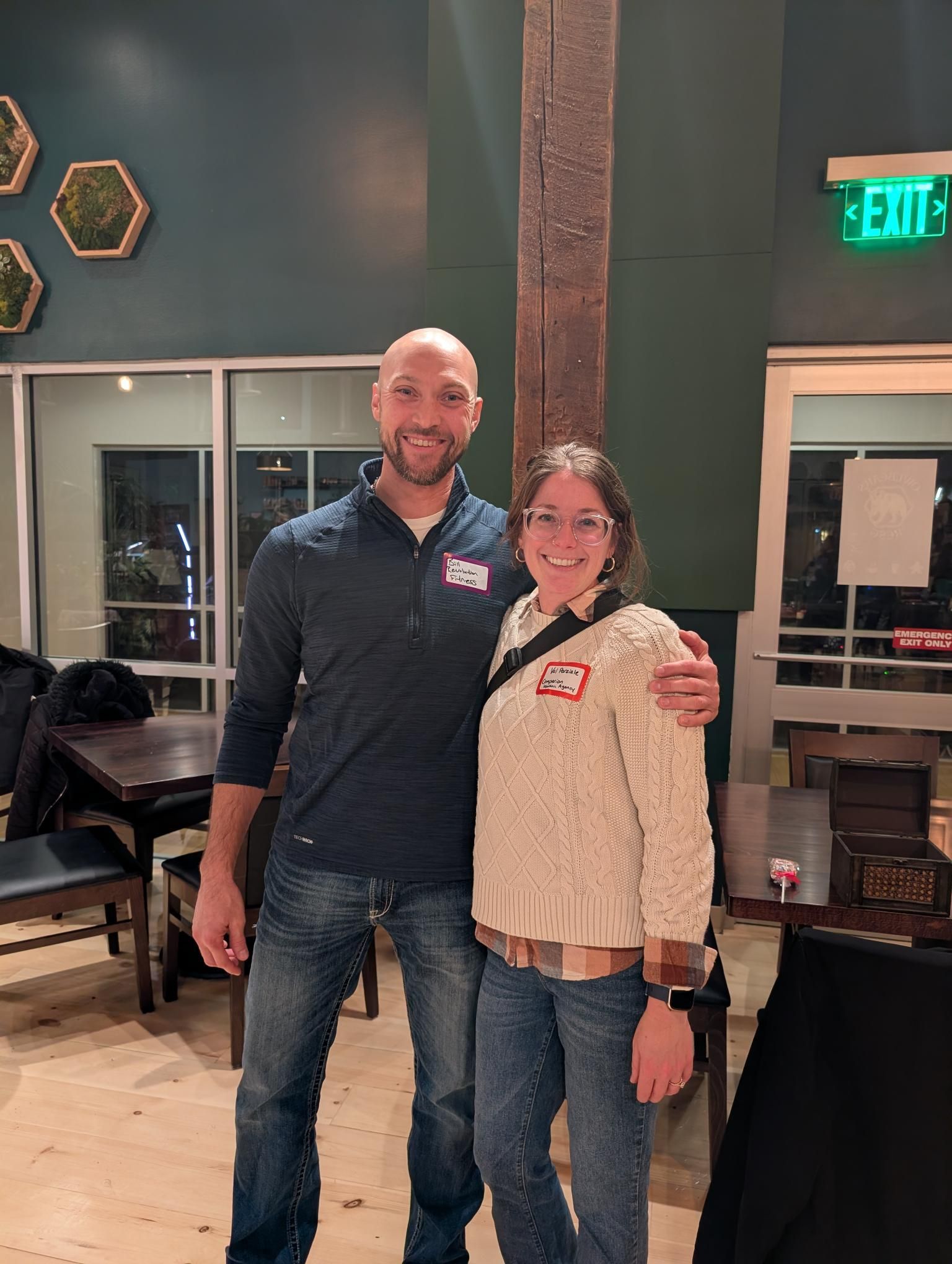 A man with a bald head and a woman wearing glasses pose for a photo. They stand indoors near a wooden support beam.