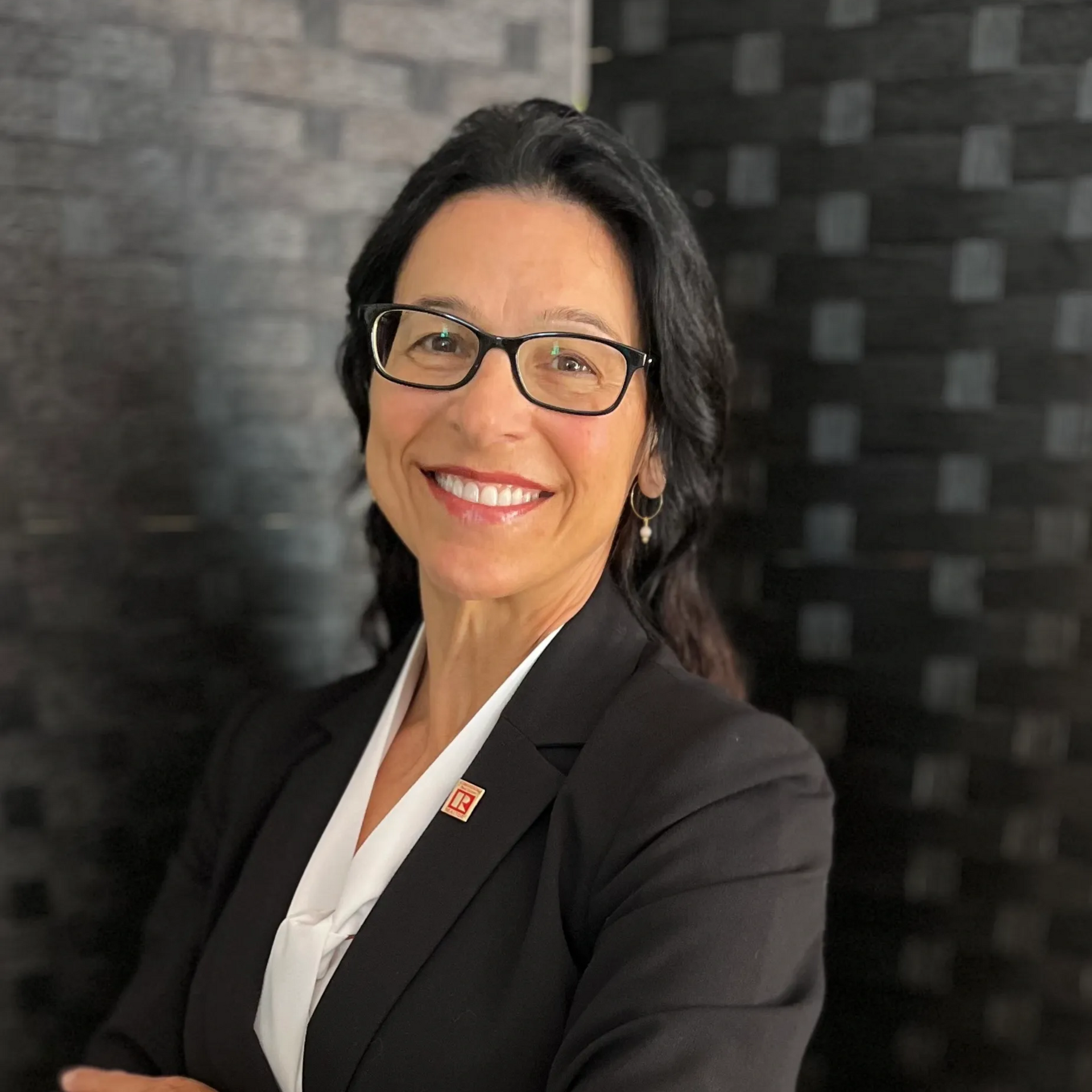 Woman with blonde hair, smiling, wearing a white shirt and blue pinstripe blazer, in a natural outdoor setting.