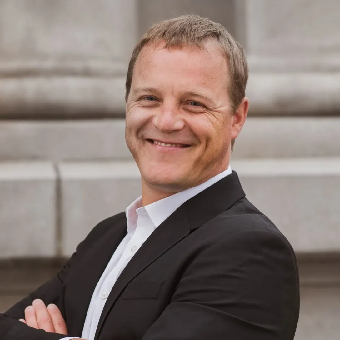 Man with short dark hair, goatee, and black tie smiles at the camera in front of a blurred outdoor backdrop.