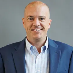 Bald man in blue suit and light blue button-down shirt smiling, set against a gray background.