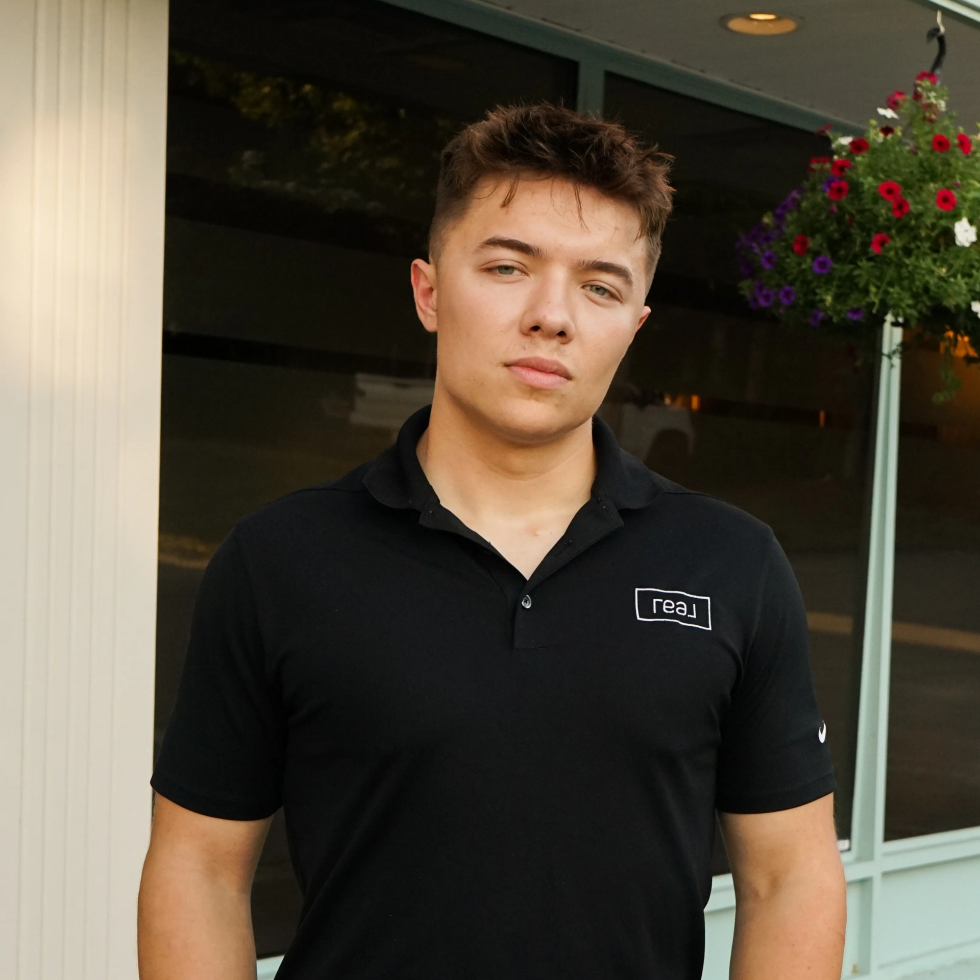 Man in black polo shirt stands in front of a window, looking directly at the camera.