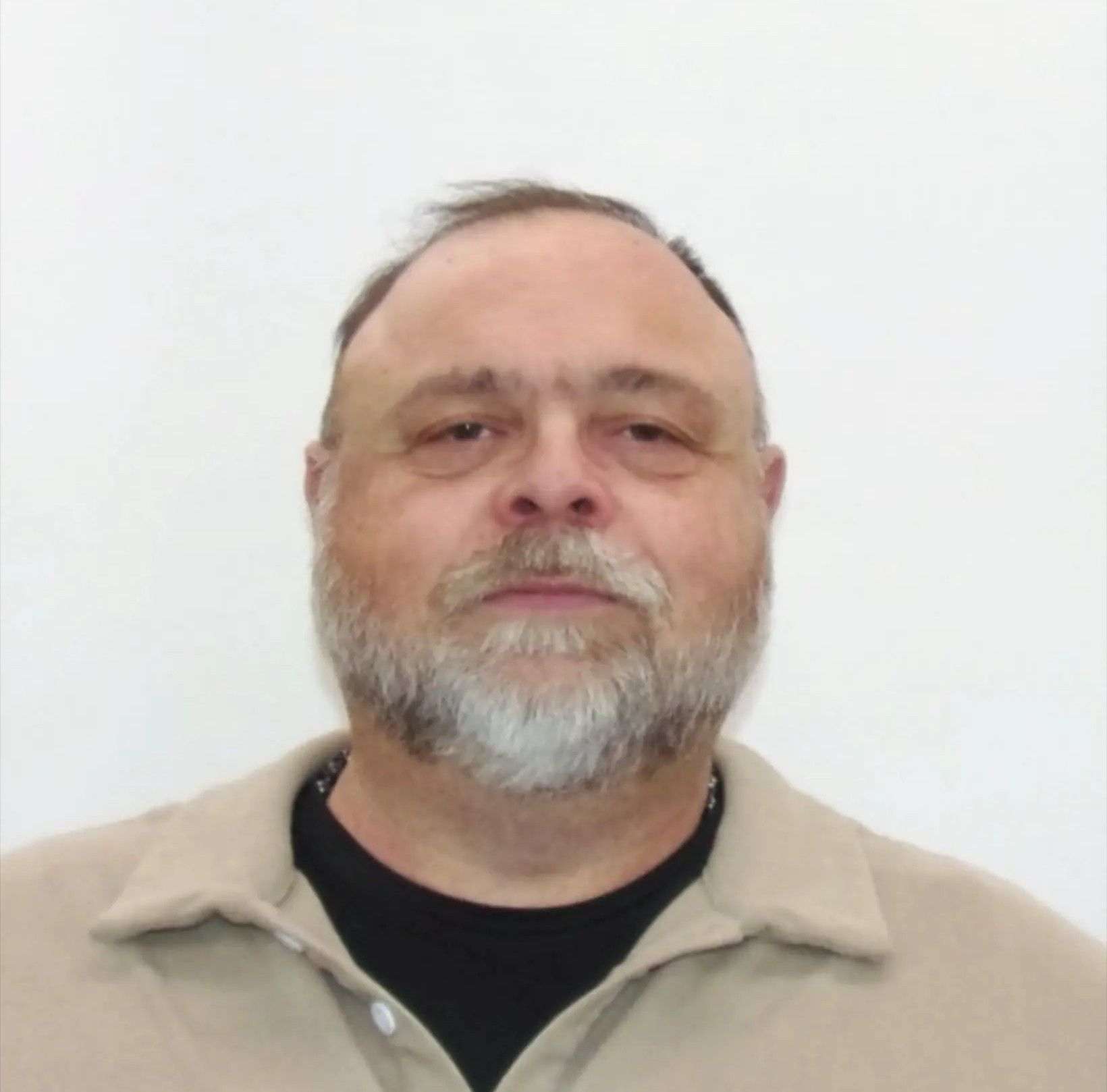 A person with a grey beard wearing a tan polo shirt over a black shirt against a plain white background.