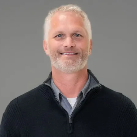 Man with graying hair and beard smiling, wearing a dark sweater over a gray collared shirt, against a gray background.
