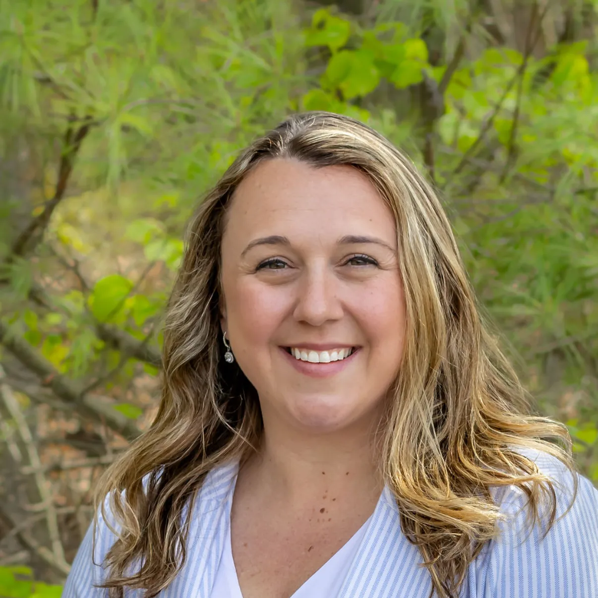 Woman with blonde hair, smiling, wearing a white shirt and blue pinstripe blazer, in a natural outdoor setting.