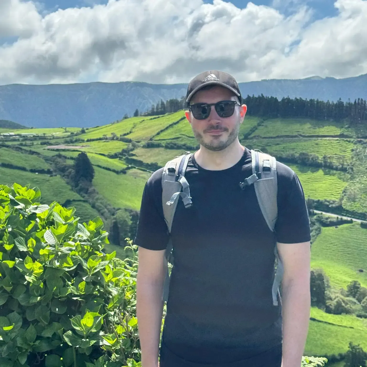 Man in sunglasses and hat with backpack, stands in front of green hills.