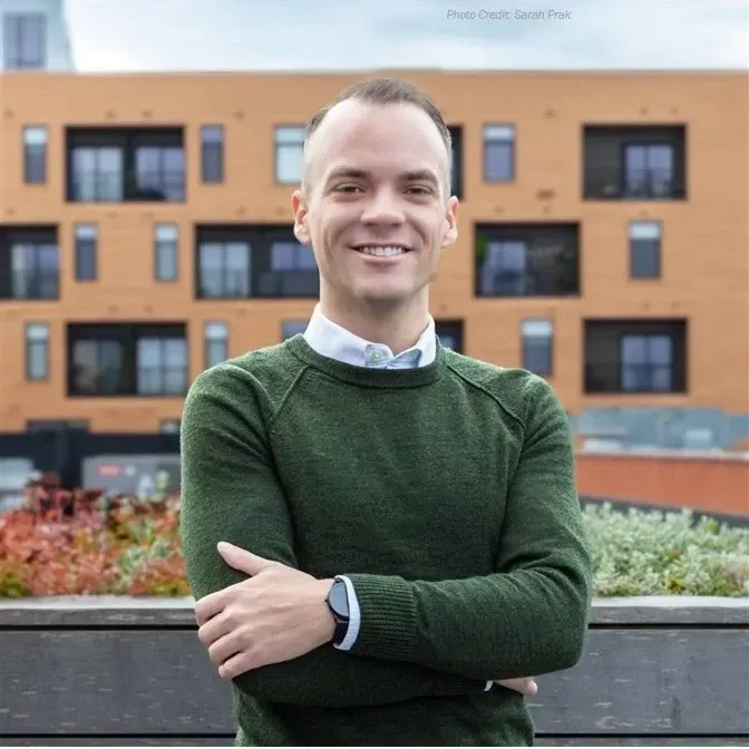 A smiling man in a green sweater poses outdoors with arms crossed, in front of a multi-story building.