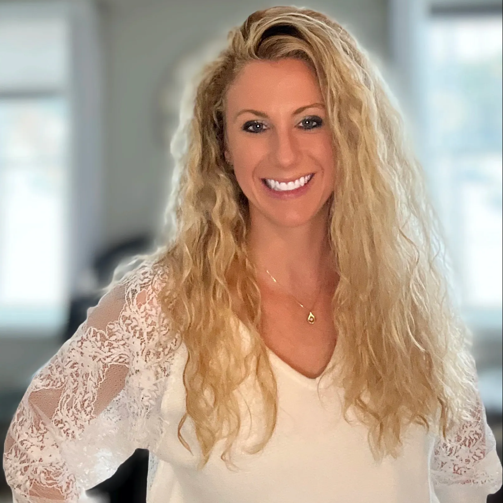 Woman with long, wavy blonde hair, smiling, wearing a white lace-sleeved shirt, in a room.