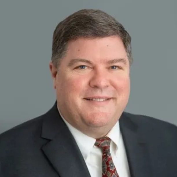 Man in a suit and tie smiles at the camera against a gray background.