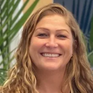 Woman in blazer smiles, rests hand on chin, wearing glasses on head, white background.