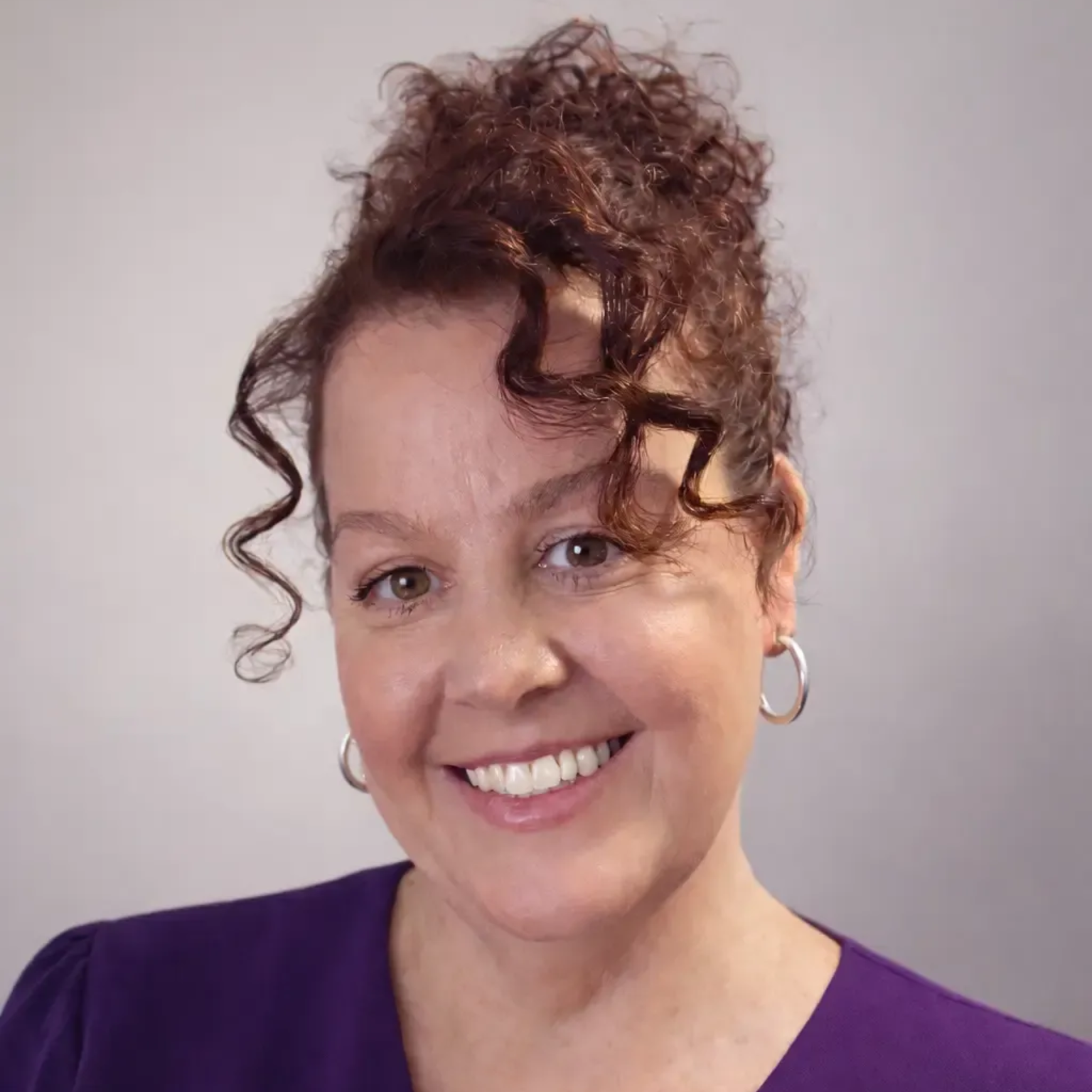 Smiling person with curly updo hair wearing a purple top against a light gray background