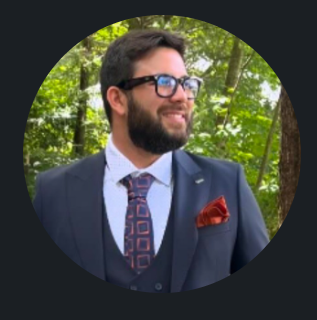 Man with short curly gray hair, wearing earbuds, looking directly at the camera. He wears a dark blue jacket.