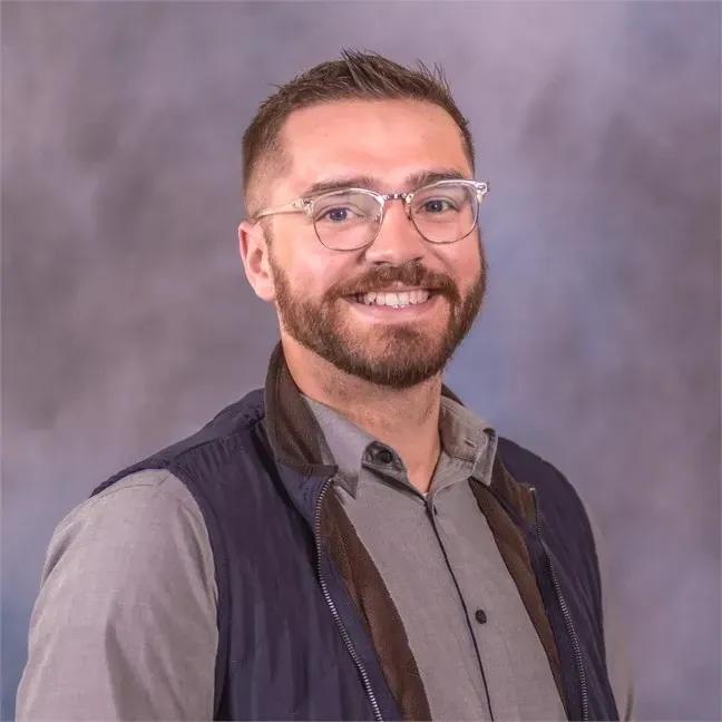 Man with glasses and a beard smiling, wearing a gray shirt and blue vest, against a blurred blue background.