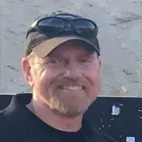 Man with beard and sunglasses on cap smiles.