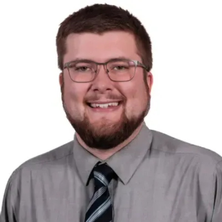 Man with glasses, beard, and tie, smiling, wearing gray shirt.