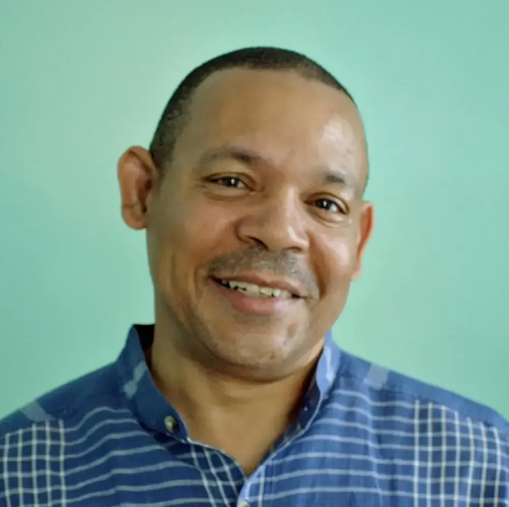Man with shaved head smiling, wearing a blue plaid shirt, against a light green background.