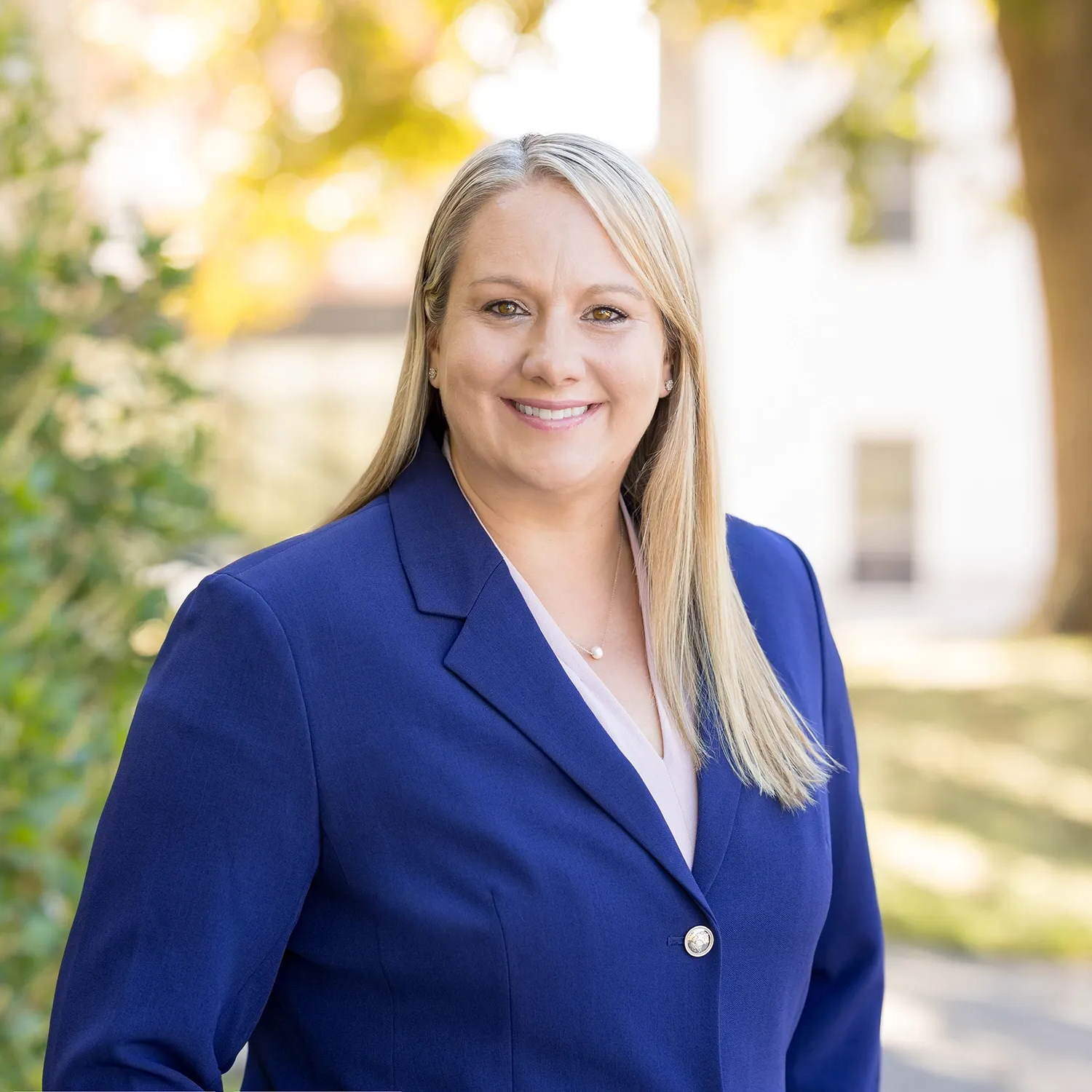 Woman in a blue blazer smiles, standing outdoors in front of trees and a building.