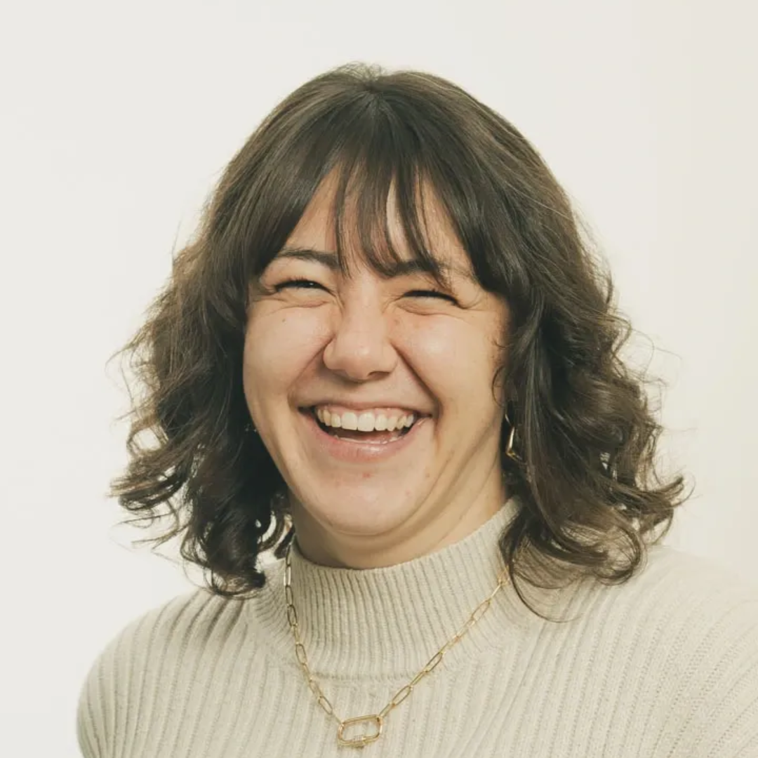 Smiling person with curly dark hair in a cream sweater against a light background. Taylor Blackdale