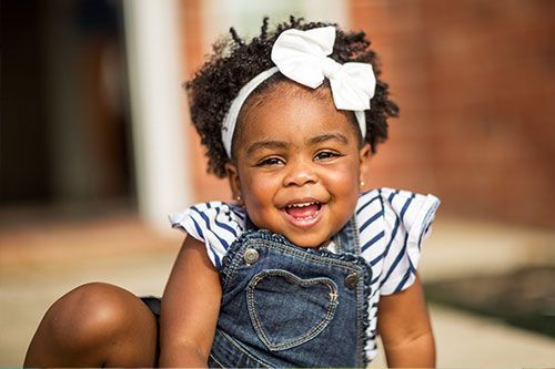 Smiling child wearing a white bow headband, denim overalls, and striped shirt outdoors.