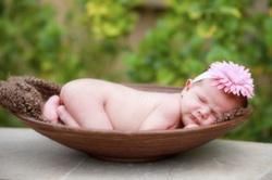 Newborn baby sleeping in a brown bowl, wearing a pink flower headband, outdoors.