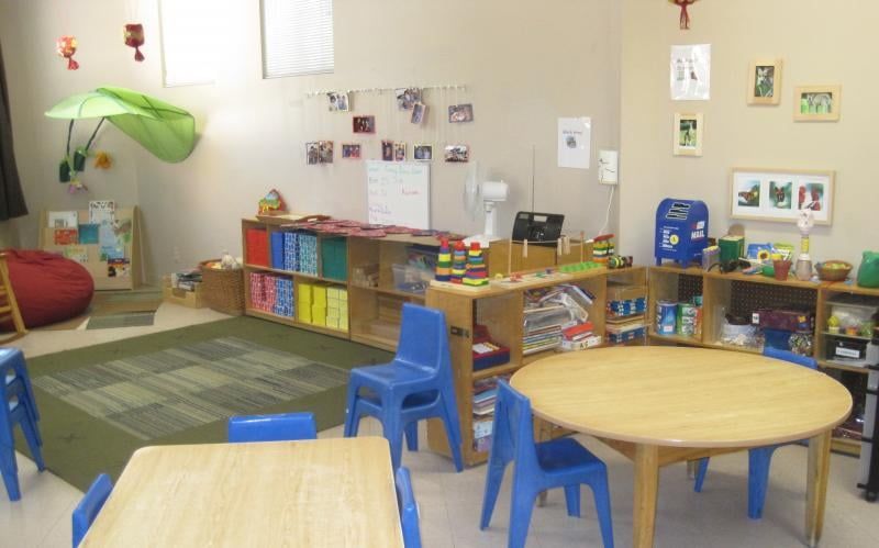 Preschool classroom with blue chairs, wooden tables, shelves with toys, and artwork on the walls.