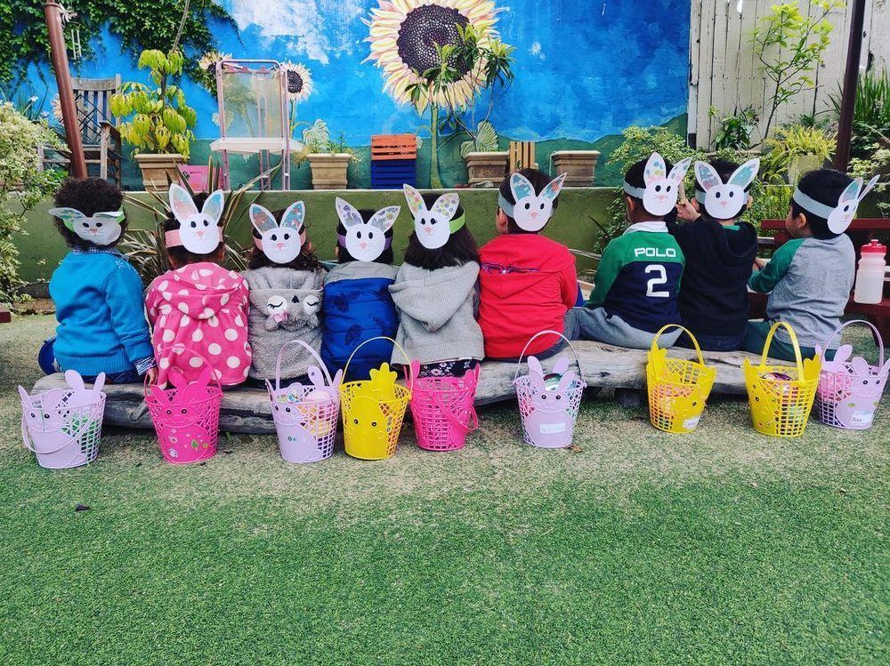 Children wearing bunny masks, sitting in a row with Easter baskets in front of a colorful backdrop.