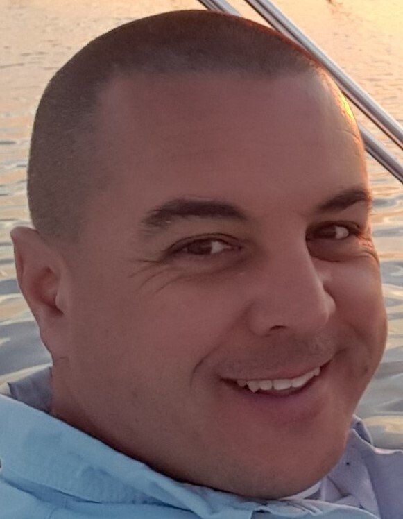 Man with short hair, smiling, close-up shot. Outdoors, light blue shirt, boat in background.
