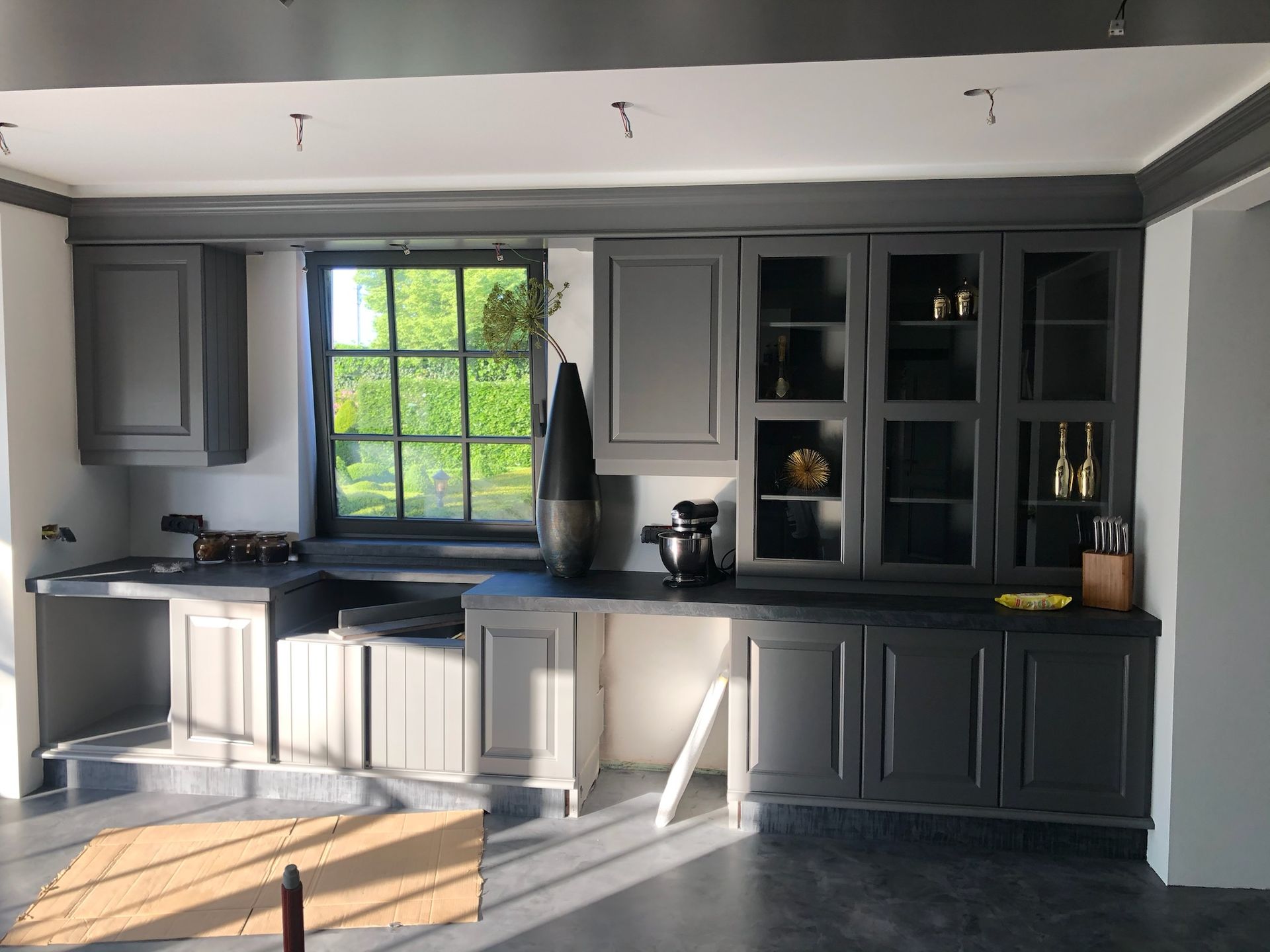 Grijze keukenkastjes met zwarte aanrechtbladen, raam met groen en donkere afwerking.