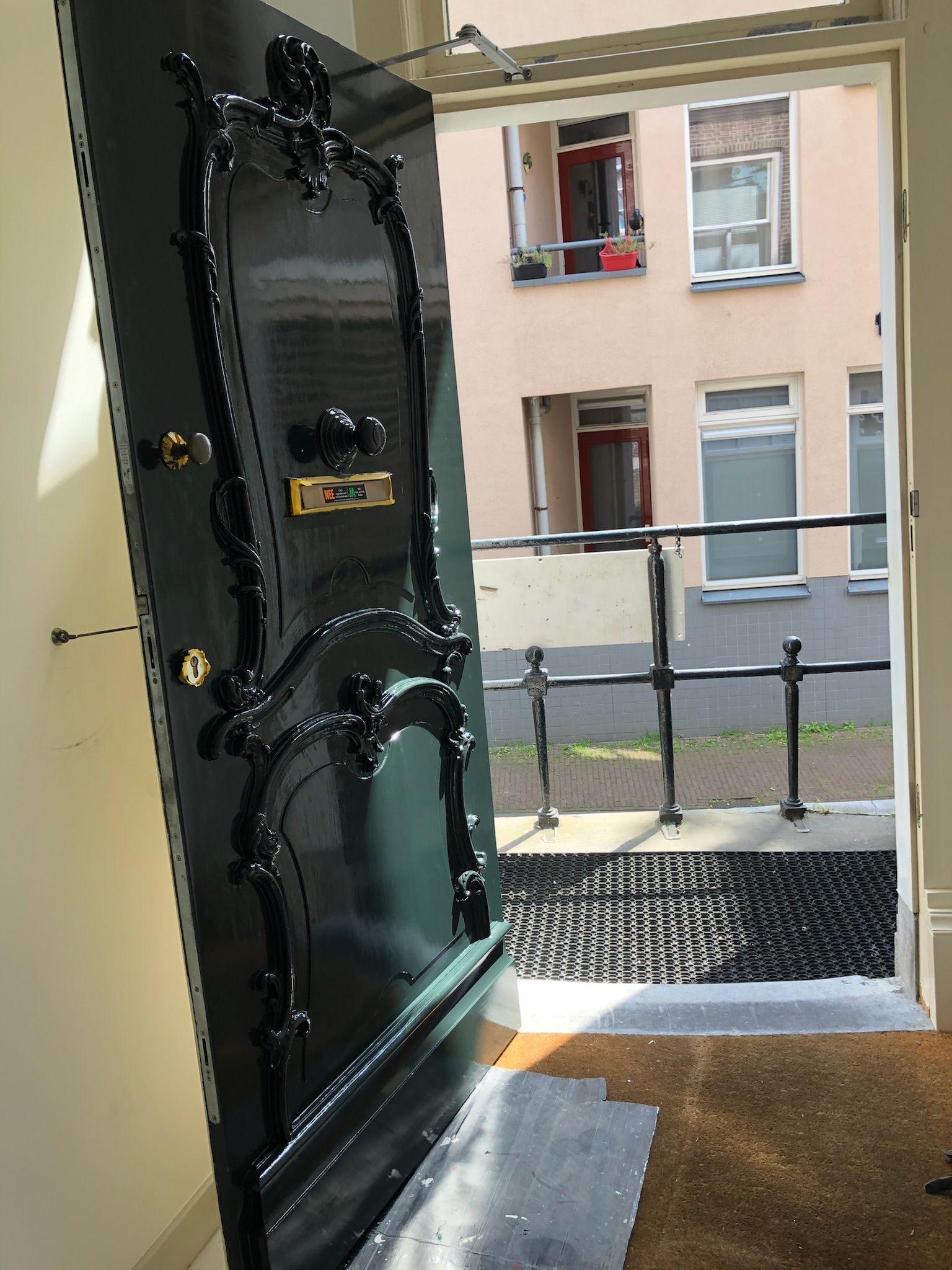 Versierde, zwarte deur staat op een kier, waardoor de buitenkant met het gebouw en de leuning te zien is.