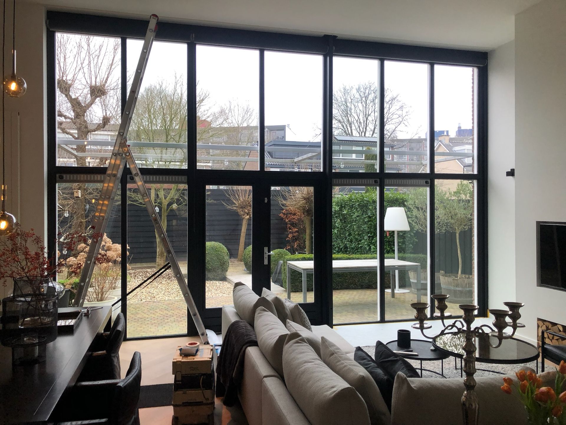 Interieur van een moderne woonkamer met grote ramen die uitkijken op een tuin. Zwarte kozijnen, neutraal meubilair.