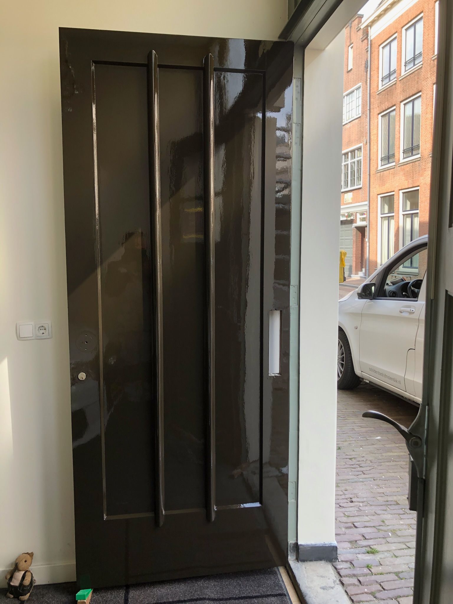 Een donkerbruine voordeur staat op een kier en aan de overkant van de straat zijn een witte auto en een bakstenen gebouw te zien.