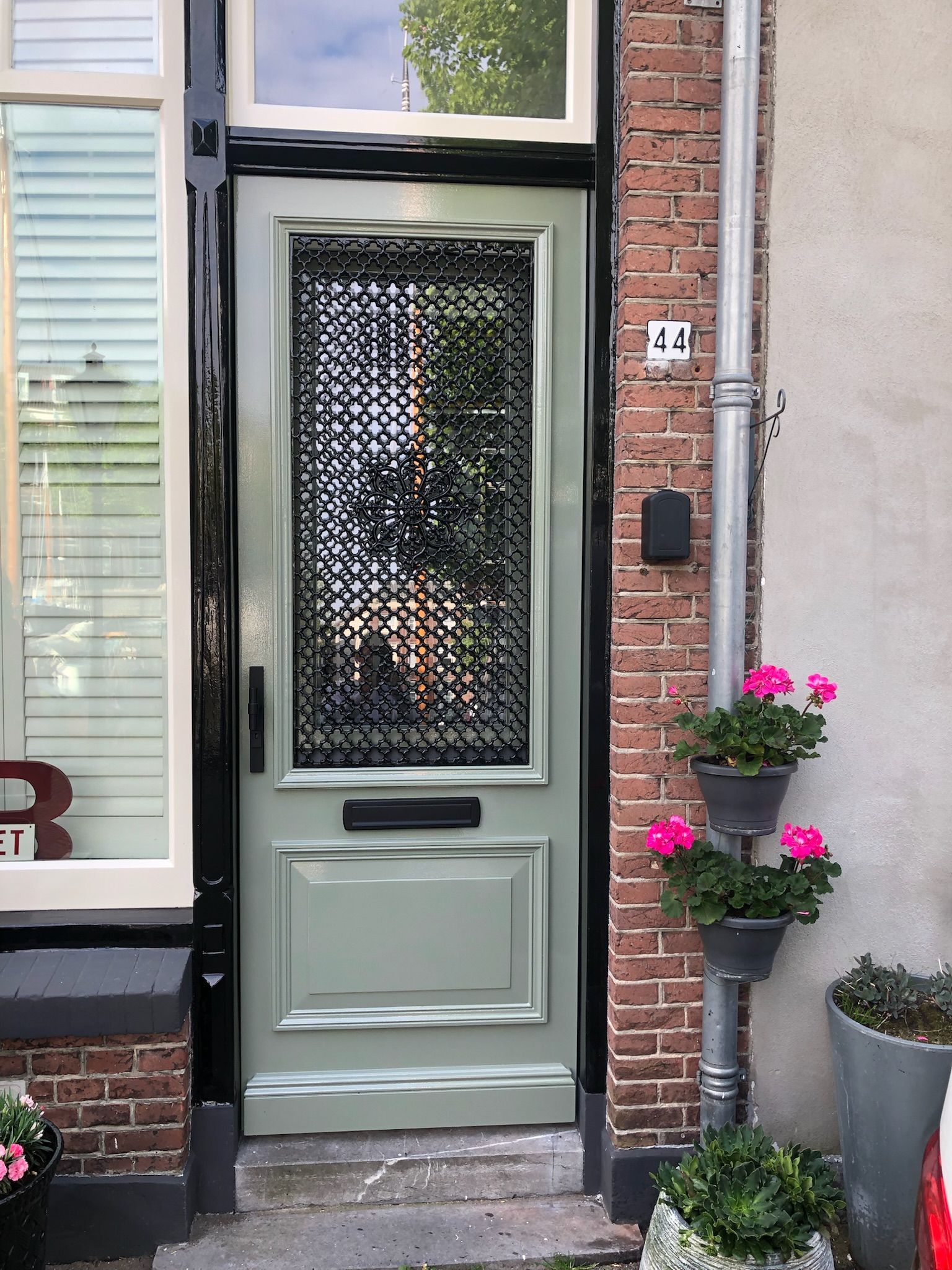 Groene voordeur met decoratief zwart traliewerk, geflankeerd door bakstenen en potplanten; nummer 44.