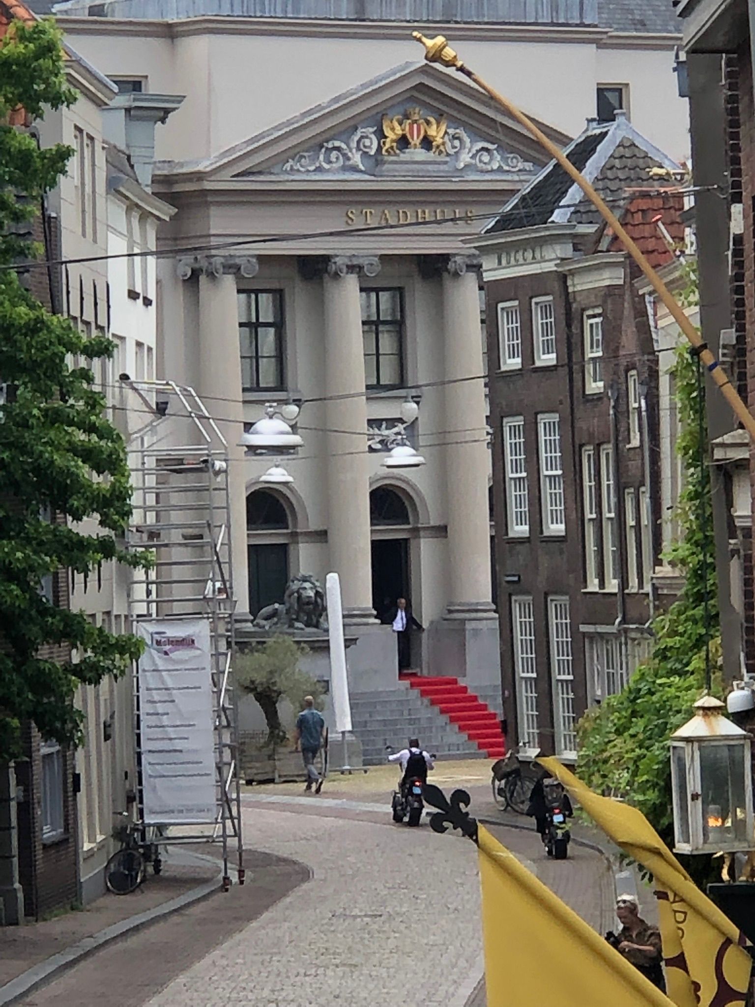 Een geplaveide straat leidt naar een klassiek gebouw met zuilen, een rode loper en mensen.