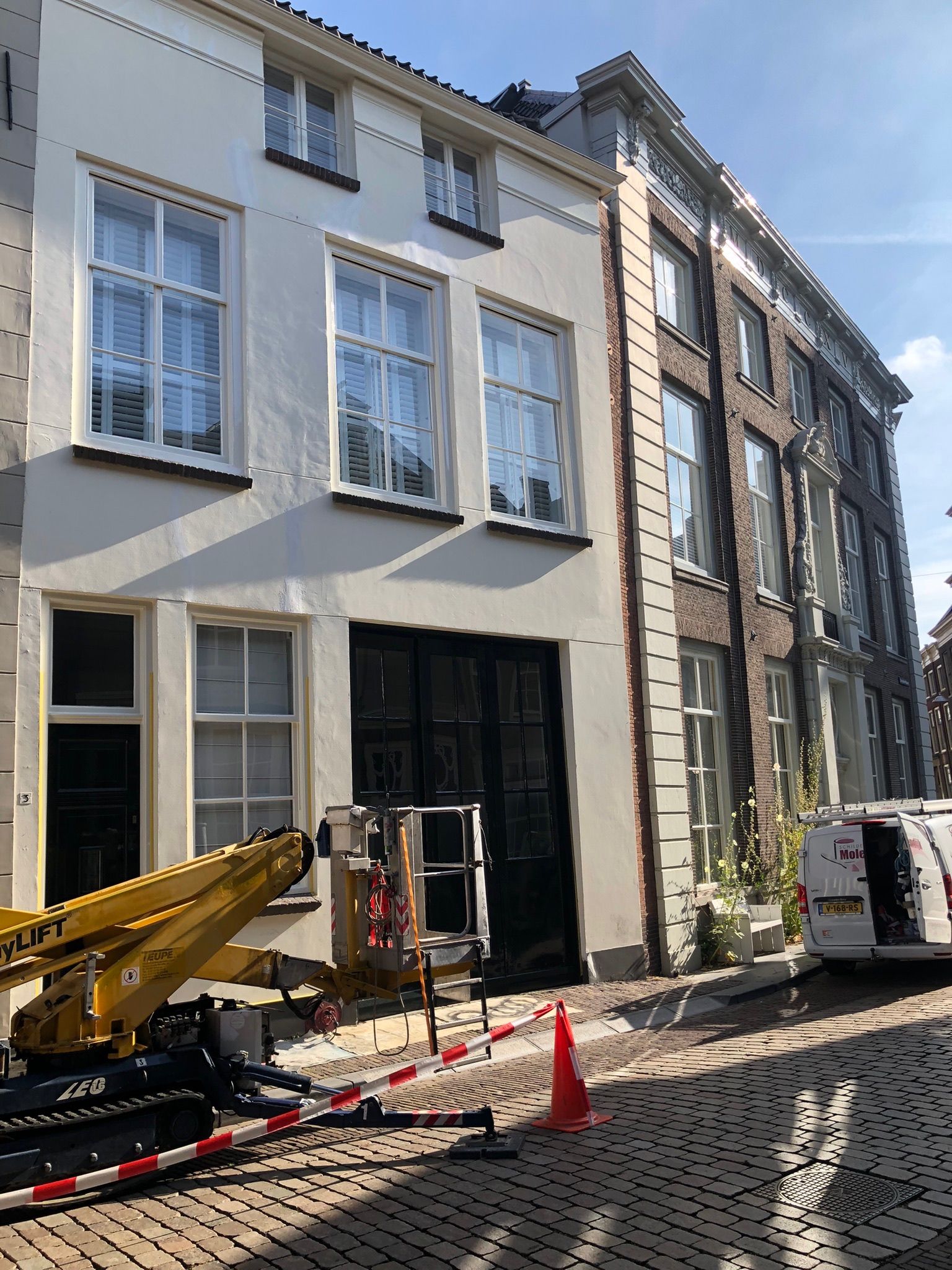 Een verweerde, witgeschilderde houten deur met zwarte scharnieren en versieringen, die een klein beetje openstaat, in een lichtgekleurde kamer.