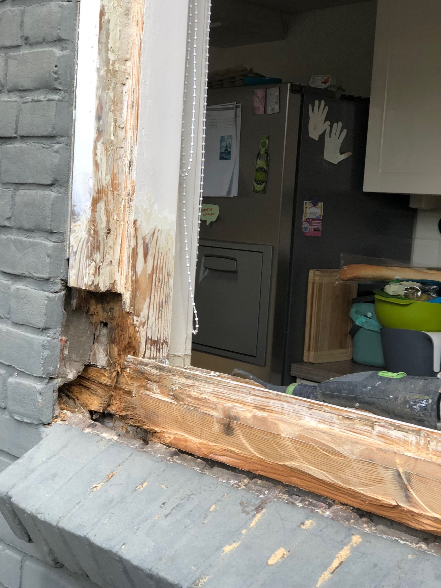 Beschadigd raamkozijn, met houtrot en bakstenen aan de buitenkant, uitzicht op een keuken.