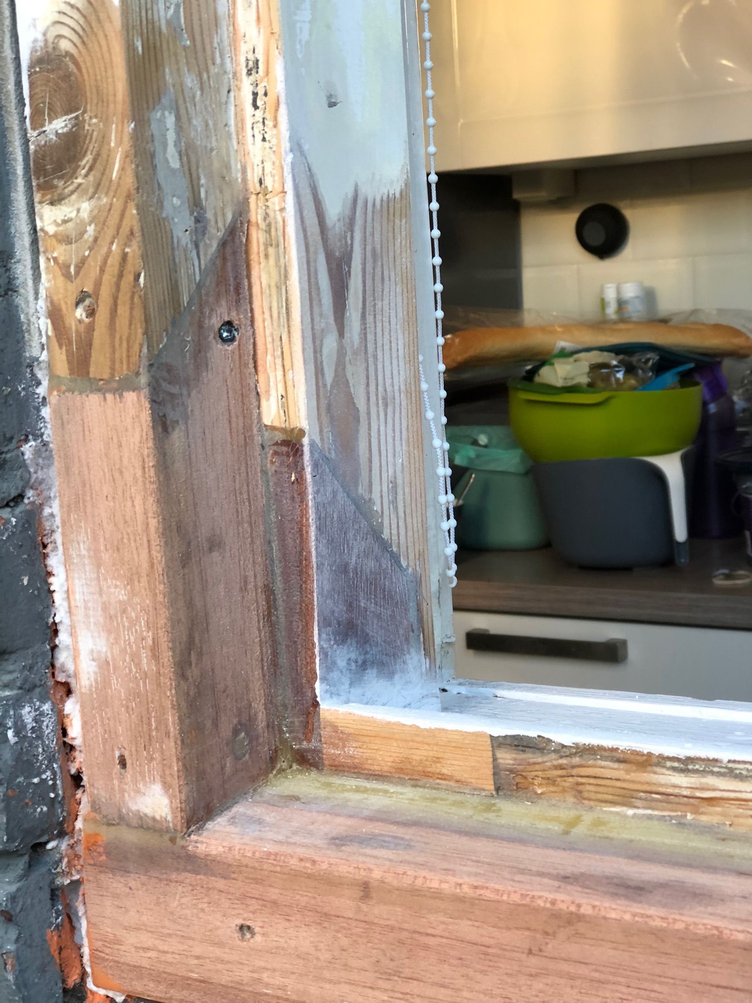 Houten raamkozijn met afbladderende verf en gaas; op de achtergrond is de keuken zichtbaar.