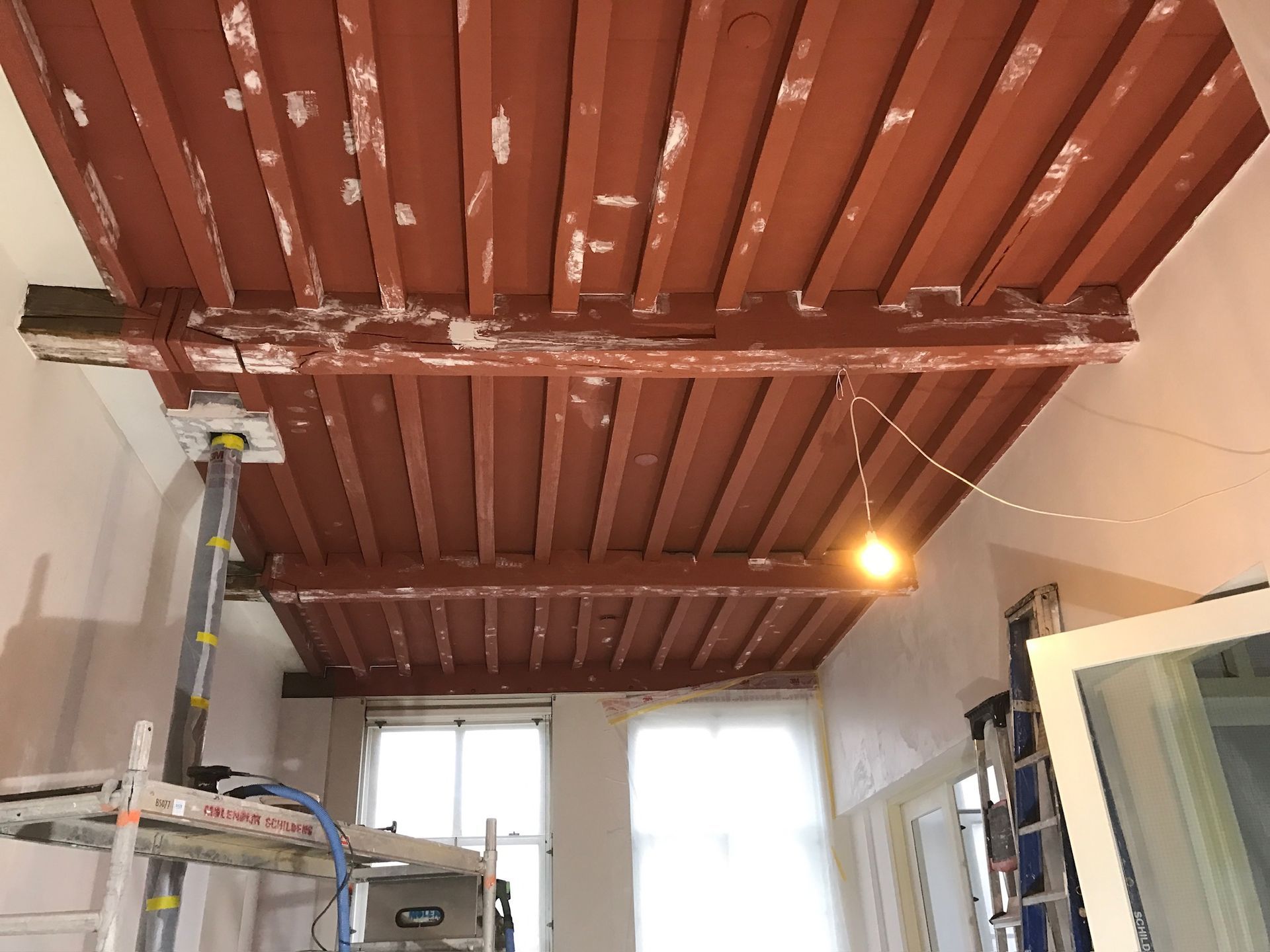 Rood geverfde houten plafondbalken in een in aanbouw zijnde ruimte met een gloeilamp en steigerwerk.