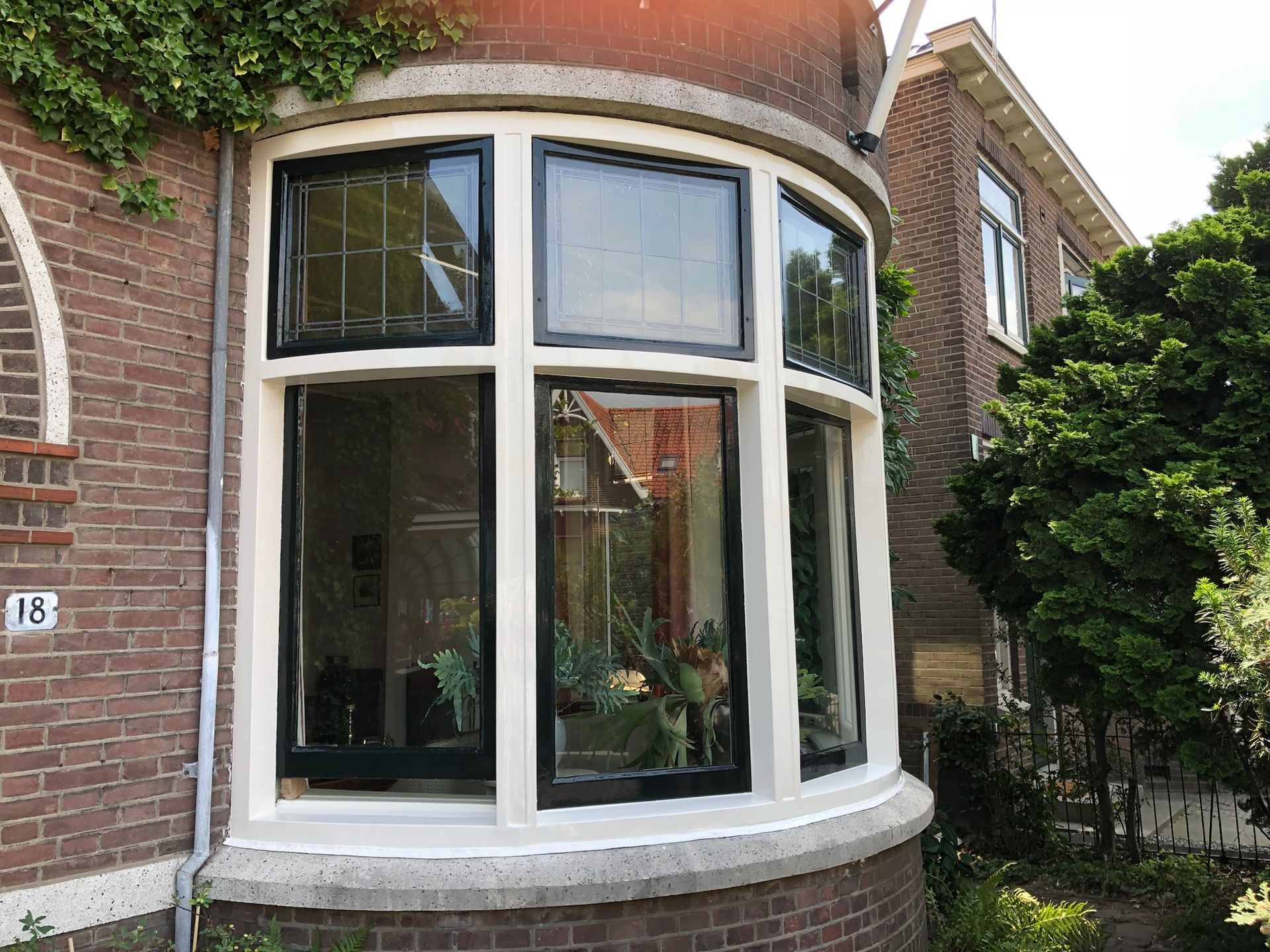 Curved window in a brick building with white frames and black borders, reflecting a house and greenery.