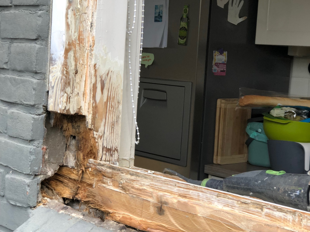 Rotting window frame, partially exposed, next to painted brick wall and interior view with metal cabinet.