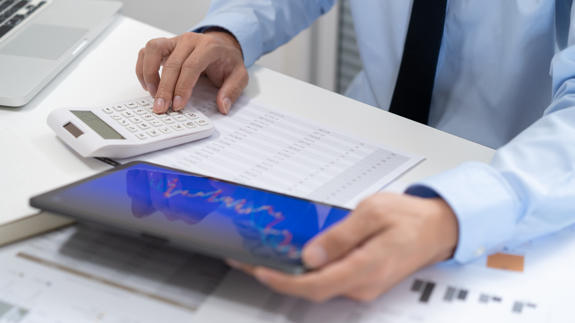 Person in a blue shirt uses calculator, holding a tablet with a financial chart, working at a desk.