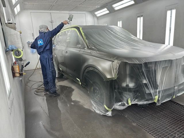 A worker sprays paint on a car in a paint booth, the covered car is partially exposed.