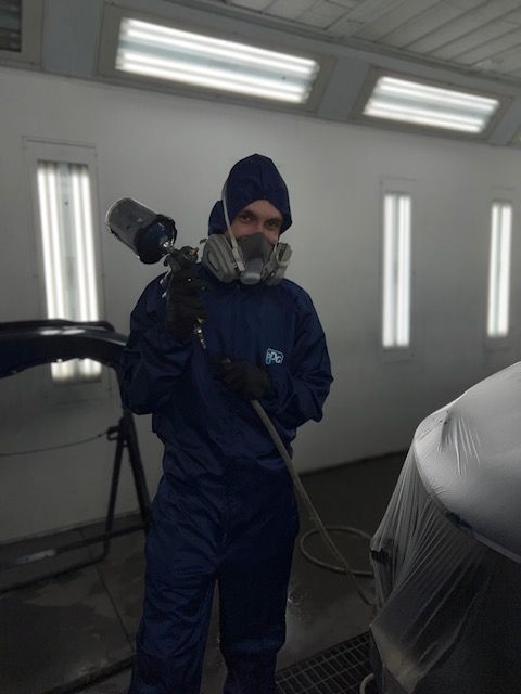 Person in blue coveralls and respirator holding a spray gun in a paint booth.