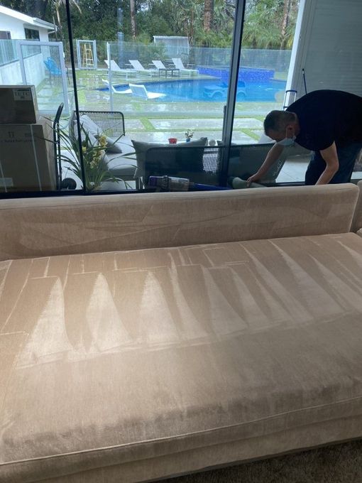 Person cleaning a beige couch in a bright living room with a pool visible through the windows