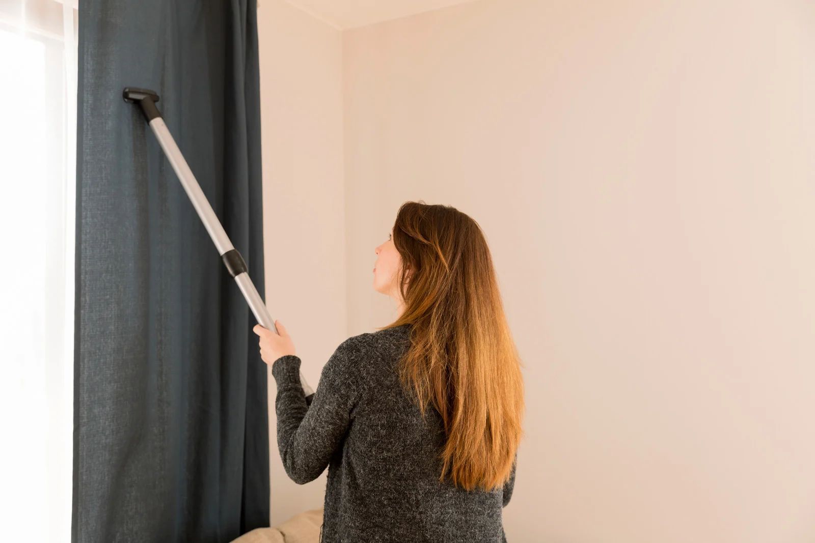 Woman vacuuming a dark blue curtain with a long vacuum attachment in a room with white walls.