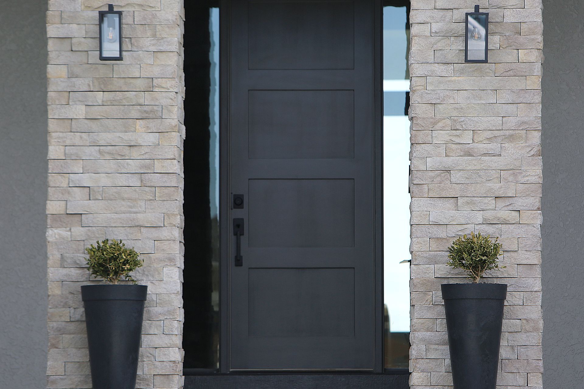 Modern grey door with rectangular panels, stone columns, potted plants, and sconces.