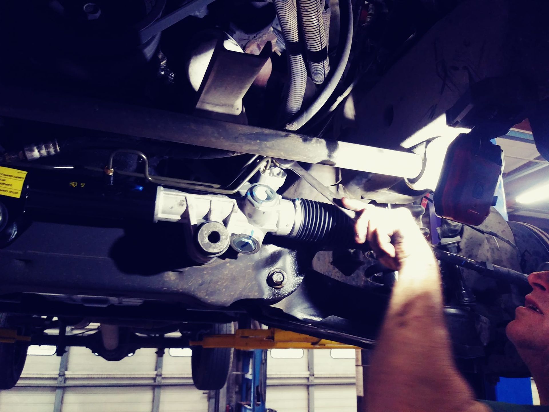 Mechanic working on the undercarriage of a vehicle, pointing at the steering rack in a repair shop.