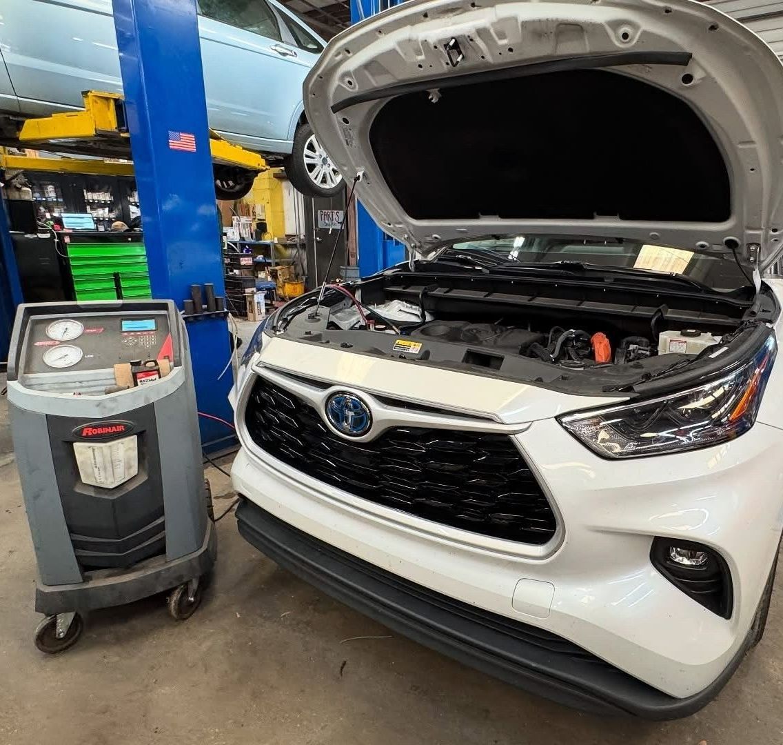 A mechanic works on a car engine with the hood open in a garage. He wears a green shirt and a hat, and there's a machine to his left.