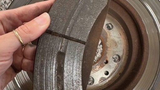 A hand holds a worn brake pad in front of a used brake rotor. The pad has a split, the rotor is grey.